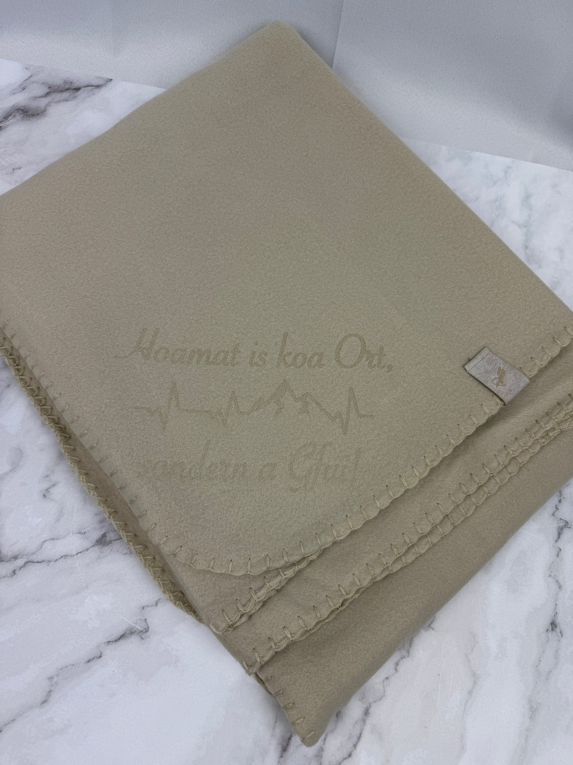 Beige Fleece-Kuscheldecke mit Aufdruck „Hoamt is koa Ort, sondern a Gfui“, gefaltet auf hellem Untergrund, weich und gemütlich