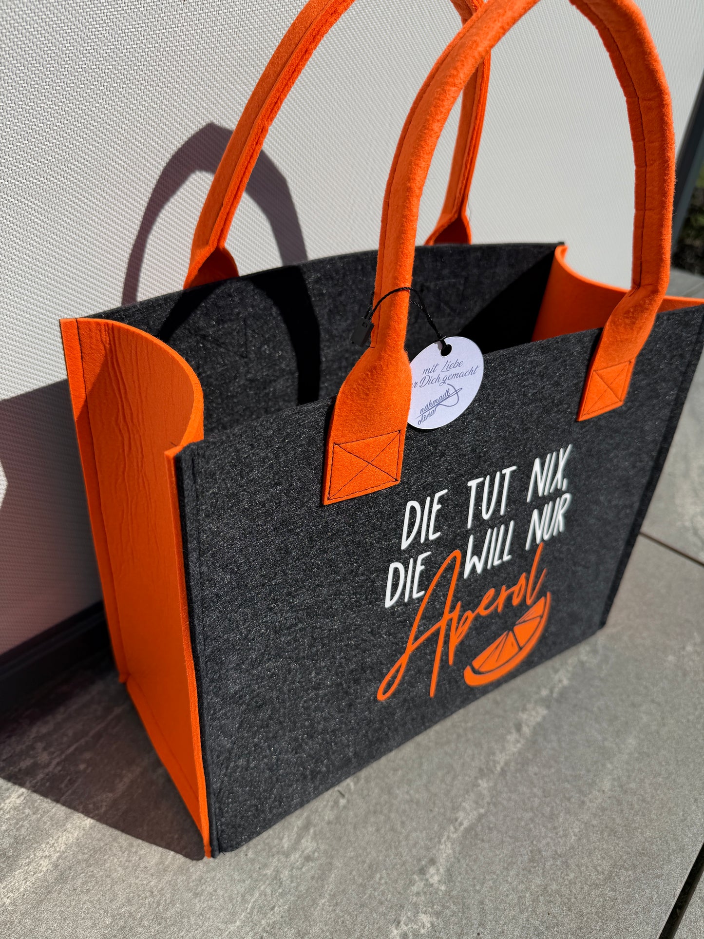 Handgefertigte anthrazitfarbene Filztasche mit orangenen Tragegriffen und dem Spruch „Die tut nix, die will nur Aperol.“ – die perfekte Tasche für Camping und Sommer.